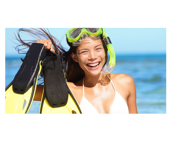 A lady standing on a beach. She is wearing goggles and a snorkel pushed up on her head and carrying flippers in her hand.